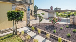 Un edificio de dos pisos con fachada de piedra, un balcón con barandillas negras y un pequeño jardín con senderos de grava y arbustos variados.