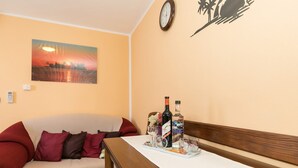 Ein gemütliches Zimmer mit einer hölzernen Bank, einem kleinen Tisch mit Tablett, auf dem eine Flasche und Gläser stehen, einem roten Sofa mit Kissen, einer Wanduhr und einem Sonnenuntergangsbild.