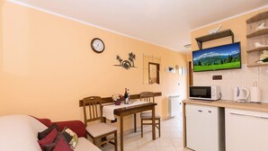 Ein gemütliches Wohnzimmer mit einem roten Sofa, einem Holzesstisch und einer Küchenzeile inklusive Mikrowelle, Wasserkocher und Fernseher, der eine schöne Landschaft zeigt.