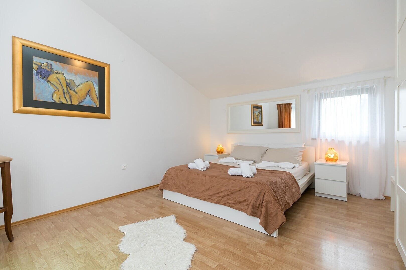 A bedroom with a bed, bedside tables, a nightstand, a painting, and a window with curtains.