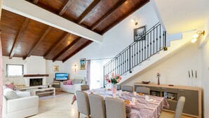 Un vaste salon doté d’un haut plafond et de poutres apparentes, un coin repas avec une table dressée pour quatre personnes, et un escalier menant à un niveau supérieur.