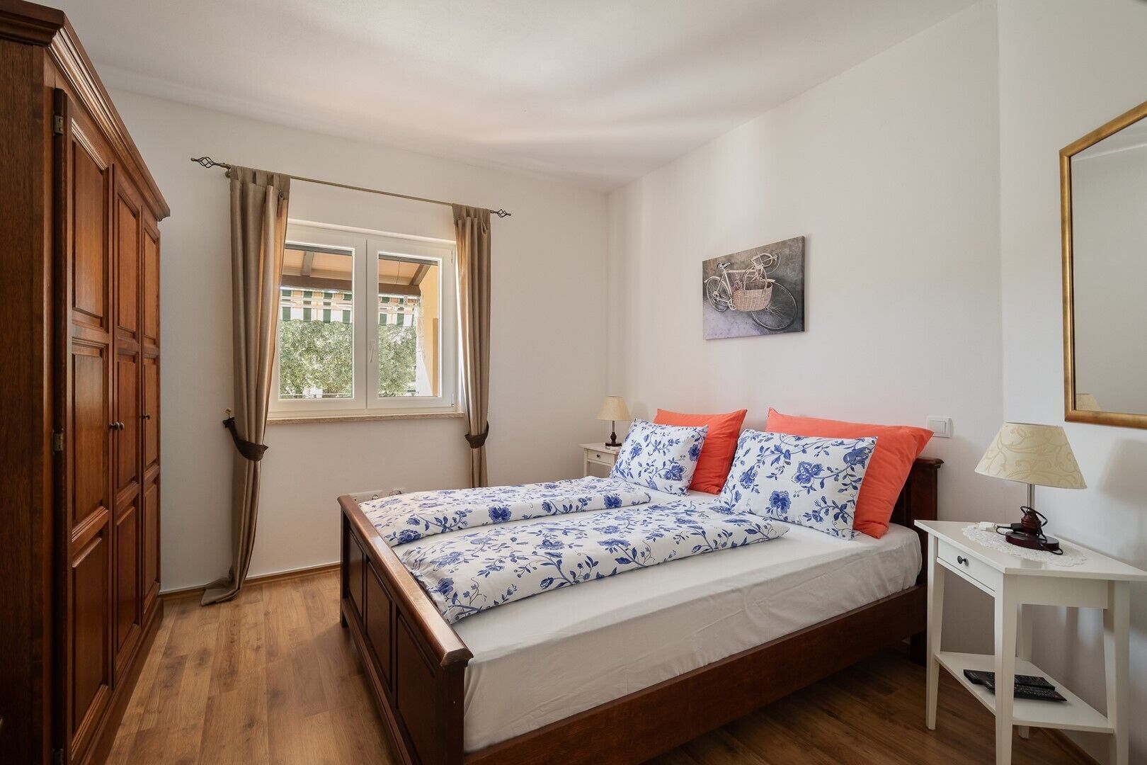 A bedroom with a wooden bed, a window with curtains, a bedside table with a lamp, and a wall-mounted artwork.