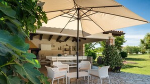 Eine Terrasse im Freien mit Steinboden, weißen Korbmöbeln und einem großen Sonnenschirm.