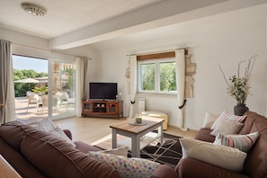 Een woonkamer met een bank, een salontafel en een televisie. Een raam met gordijnen biedt uitzicht op groen.