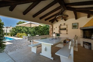 Une terrasse extérieure avec une piscine en pierre, des bancs blancs, une table et une cheminée.