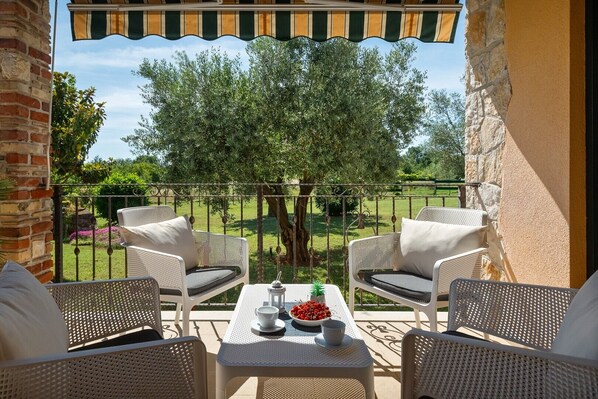 A balcony with wicker furniture, a table with a bowl of fruit, and a view of a garden with trees.