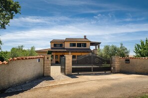 Una casa de dos pisos con entrada protegida por un portón, rodeada de un muro de ladrillo y un camino de entrada pavimentado.