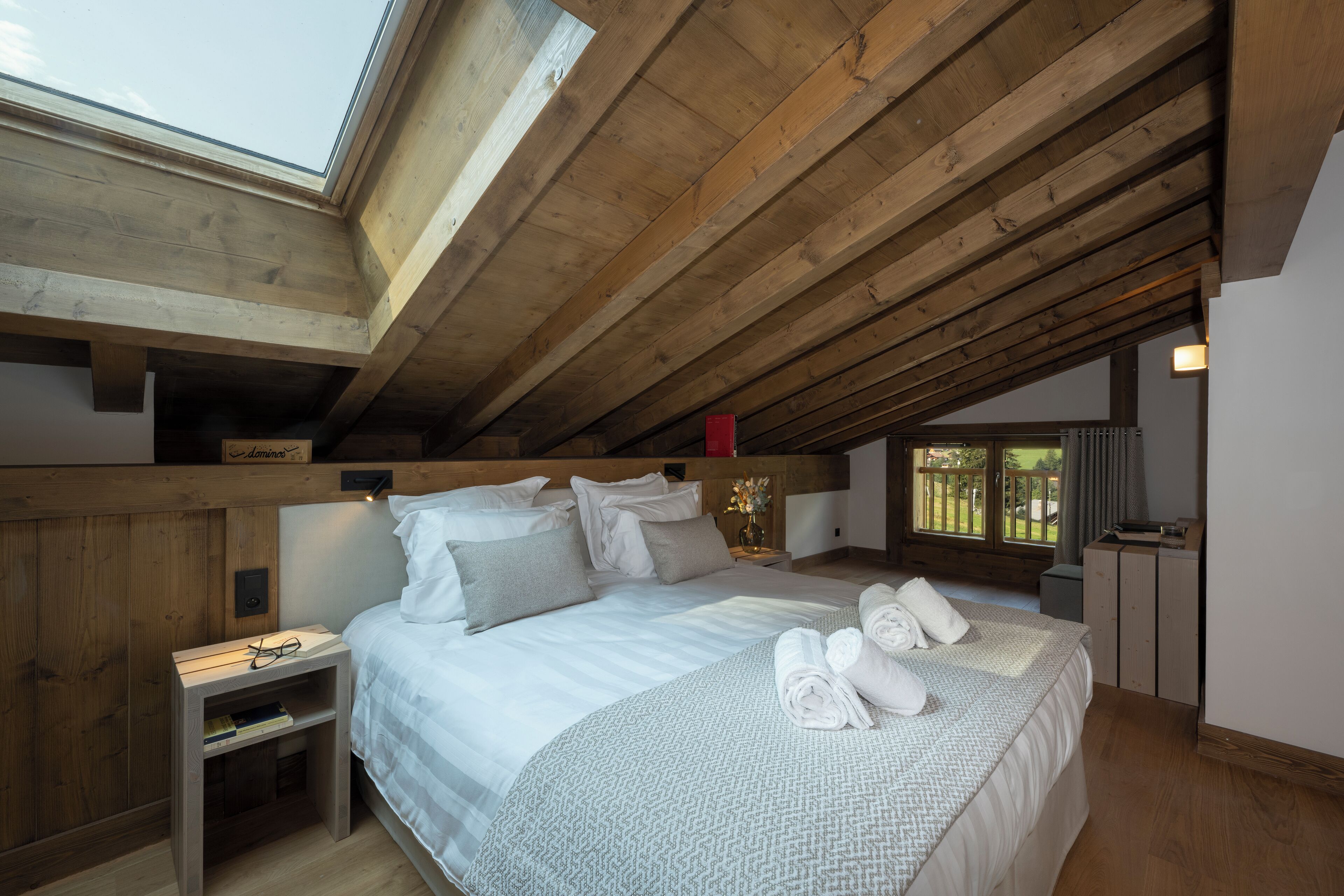 Un dormitorio con tragaluz, vigas de madera, una cama con almohadas, una mesita de noche y un buró.