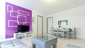 A modern living room with a glass coffee table, a flat-screen TV on a white stand, and a purple accent wall.