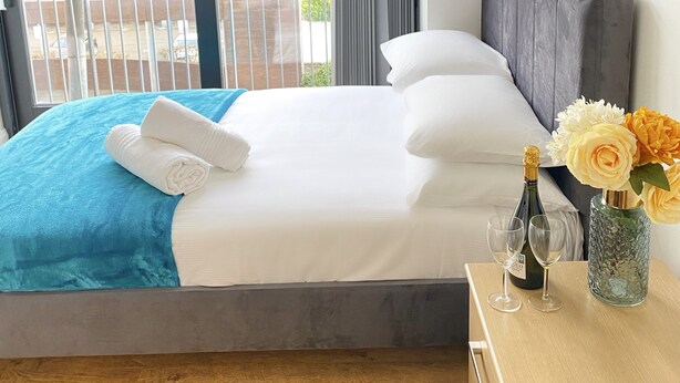 A hotel room with a bed, a nightstand, a vase with flowers, and a window with curtains. - Tudors eSuites Balcony Apartment (Birmingham)