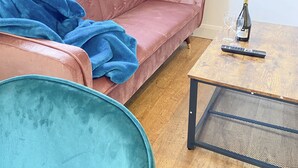 A pink sofa with a blue blanket, a teal chair, and a wooden coffee table with a bottle and glasses. - Tudors eSuites Balcony Apartment (Birmingham)
