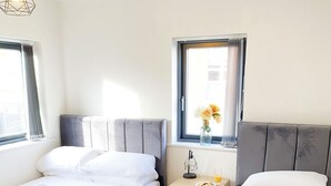 A hotel room with two beds, a nightstand, a window with curtains, and a hanging light fixture. - Tudors eSuites Balcony Apartment (Birmingham)