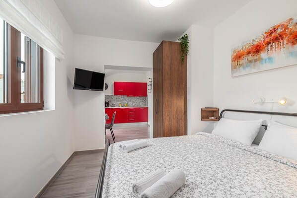 A bedroom with a bed, a TV mounted on the wall, a red kitchen in the background, and a painting on the wall. - Oroz / Oroz Studio I (Porec)