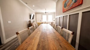 Un lungo tavolo da pranzo in legno con sedie in vimini, una ventola a soffitto e un televisore montato a parete.