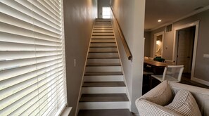 Un escalier avec une rampe, une fenêtre avec des stores et un coin repas avec une table et des chaises.