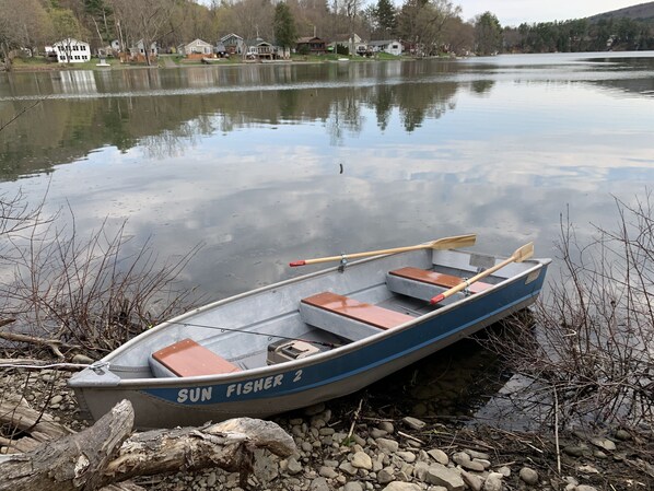 A small rowboat named 'SUN FISHER 2' with two wooden paddles, docked on a rocky shore with a calm lake in the background.
