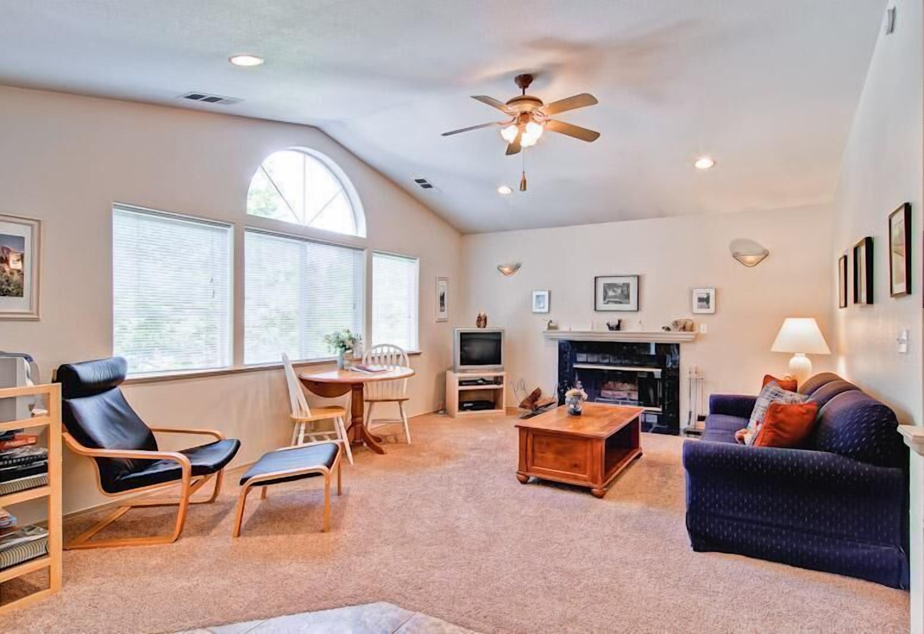 A spacious living room with a fireplace, a ceiling fan, and large windows.
