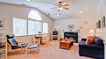 A spacious living room with a fireplace, a ceiling fan, and large windows.