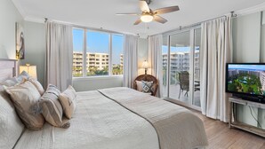 A hotel room with a large bed, a ceiling fan, a TV, and a balcony with a view of the city. - Destin West Heron 505 ~ 3BR/2.5BA ~ Bay Views ~ Overlooking the Lazy River (Fort Walton Beach)