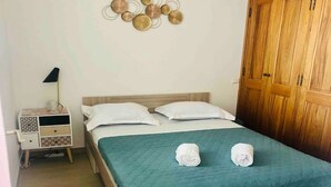 A neatly made bed with a teal bedspread and two rolled towels, a bedside lamp, a small bedside table with drawers, and a wall decoration above the bed. - Santa Lina - Magnifique T2 à 200m de la plage (Ajaccio)