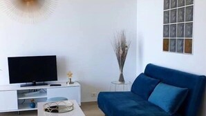 A modern living room with a flat-screen TV, a blue sofa, a coffee table, and a decorative light fixture. - Santa Lina - Magnifique T2 à 200m de la plage (Ajaccio)