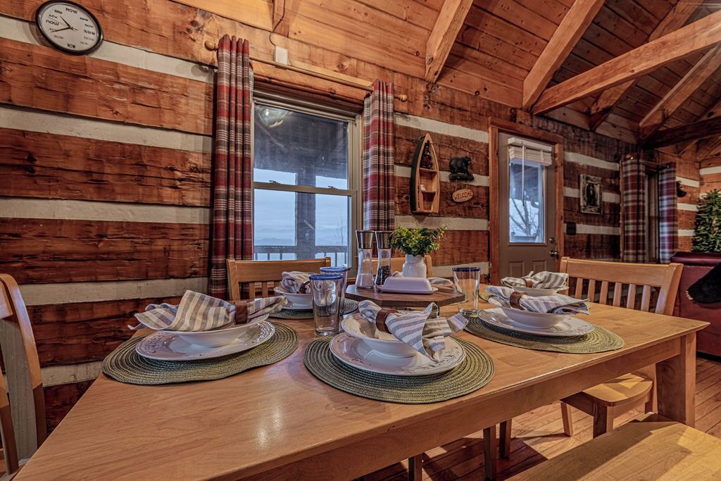 A wooden cabin interior with a dining table set for four, a clock on the wall, and plaid curtains.