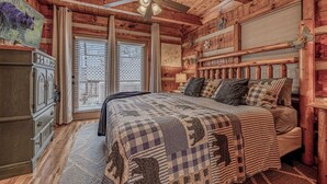 Een slaapkamer in een houten chalet, met een bed, plafondventilator en een raam met zonwering.