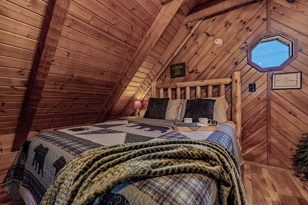 Un dormitorio de buhardilla acogedor, con techo inclinado de madera, una cama con colcha y un ventanal pequeño.