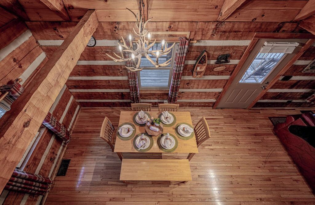 A wooden cabin interior with a dining table set for six, a chandelier, and a window with curtains.