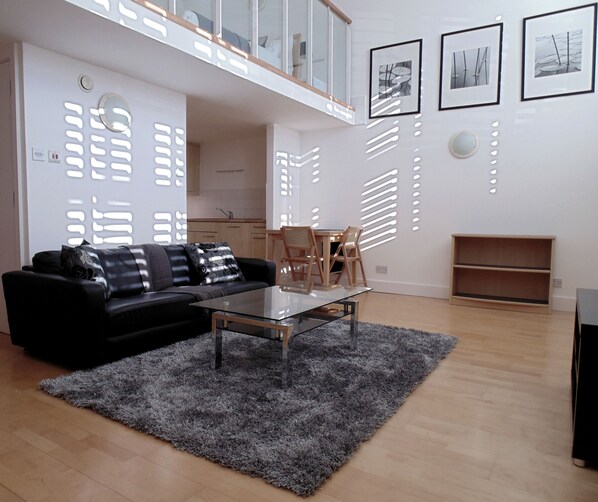 A modern living room with a black sofa, a glass coffee table, and a grey rug. There is a dining area with a wooden table and chairs. The room has white walls with decorative cutouts and three framed pictures.