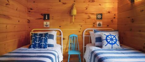 A cabin with two beds, each with a striped blue and white bedding, a blue chair, and nautical-themed wall decor.