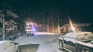 Escena nocturna con una carretera cubierta de nieve, un automóvil estacionado y un edificio con luces al fondo.
