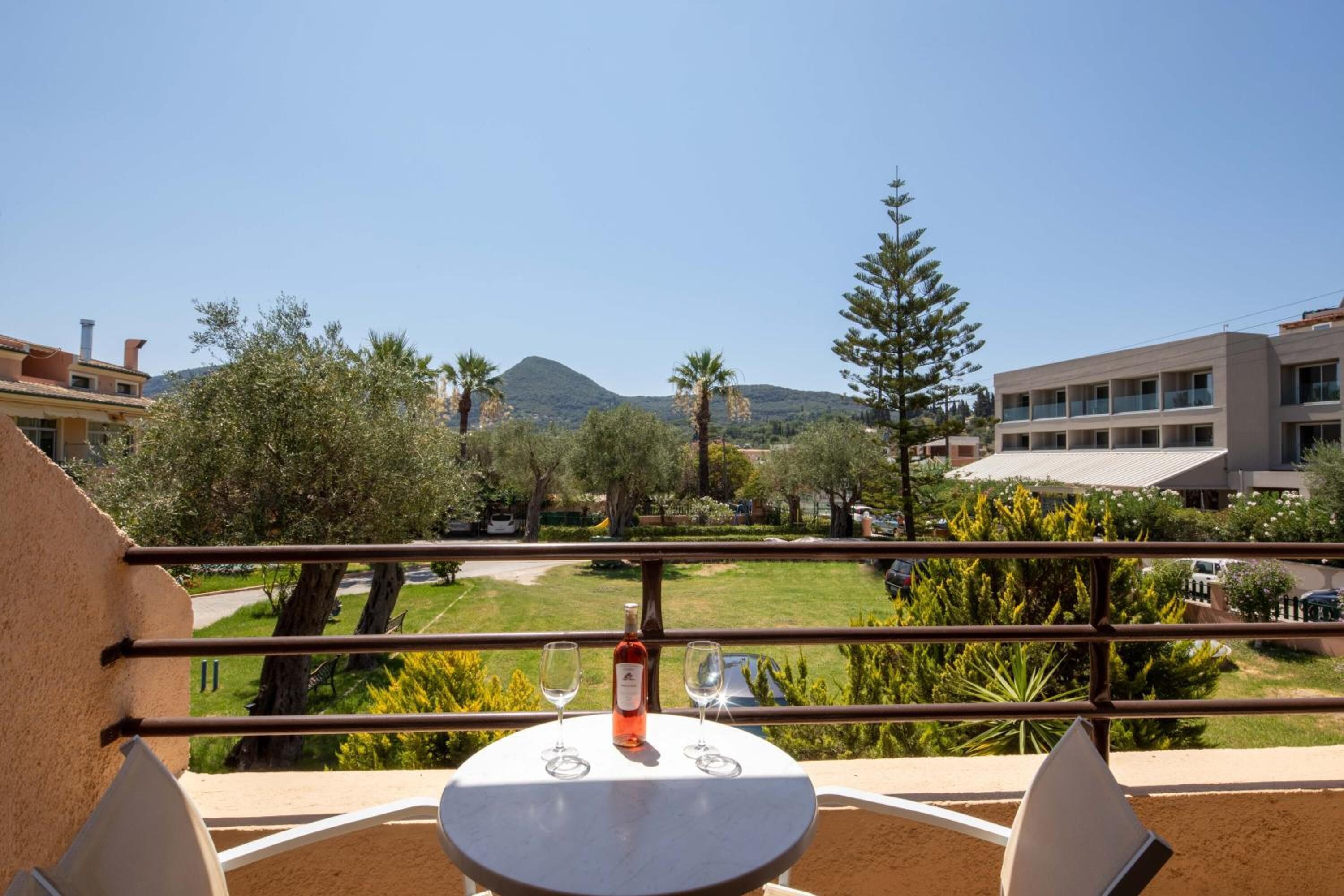 A balcony with a table and two wine glasses, overlooking a landscaped area with trees and a mountain in the background.