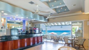 Zona de bar con ventiladores de techo, una barra con caja registradora y vista al océano a través de amplias ventanas.