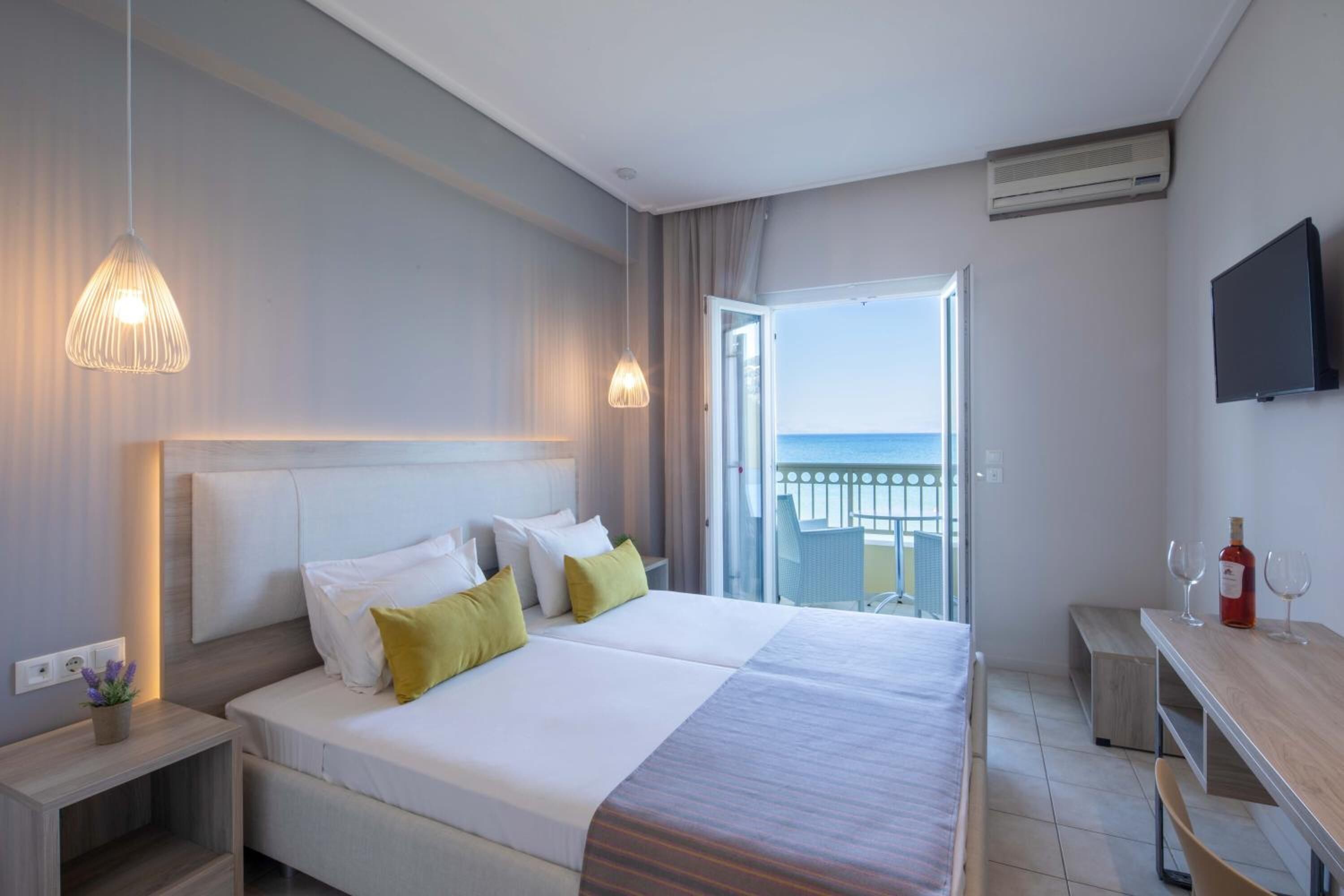 A hotel room with a large bed, bedside tables, a desk with a chair, a TV, and a balcony with a view of the sea.