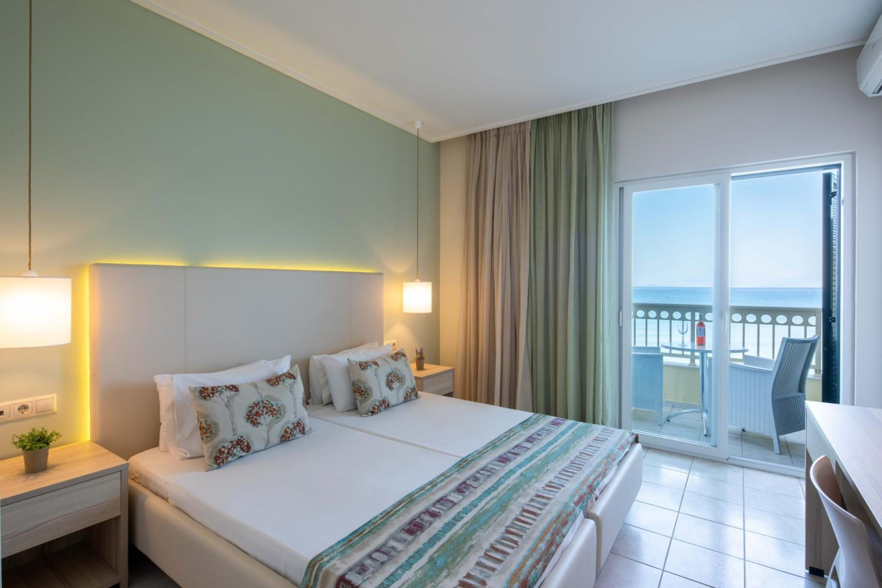 Habitación de hotel con cama, mesitas de noche, lámpara, vista al mar desde el balcón y una silla.