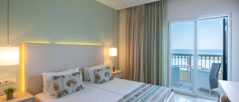 Habitación de hotel con cama, mesitas de noche, lámpara, vista al mar desde el balcón y una silla.