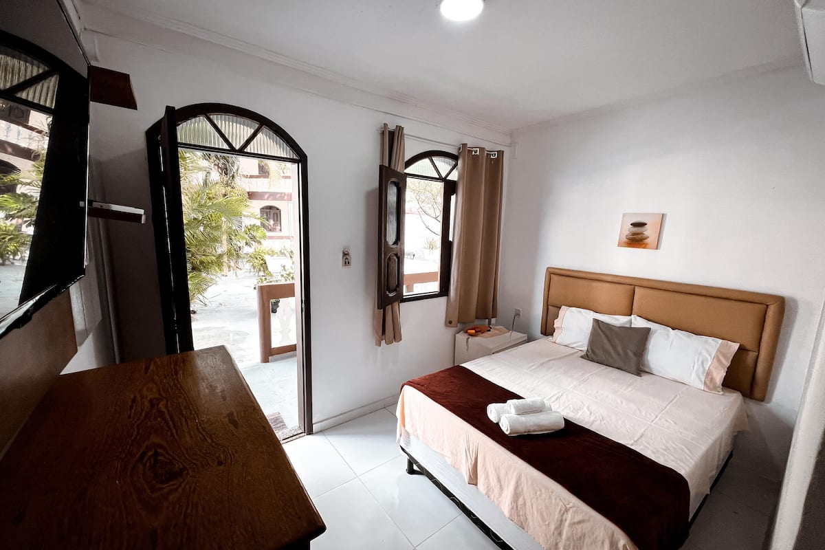 A hotel room with a bed, a wooden bench, a window with curtains, and a view of the outside.
