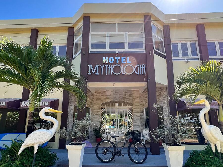 Un hotel llamado "Hotel Mythologia" con un diseño arquitectónico moderno, que presenta dos grandes estatuas de aves, una bicicleta y plantas en macetas.