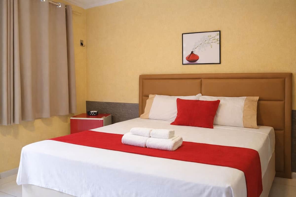 A hotel room with a bed, a nightstand, a red and white bedspread, and a framed picture on the wall.