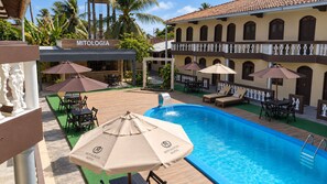 Een zwembad met ligbedden en parasols, een terras met tafels en stoelen, en een gebouw met een bord waarop 'MITOLOGIA HOTEL' staat.