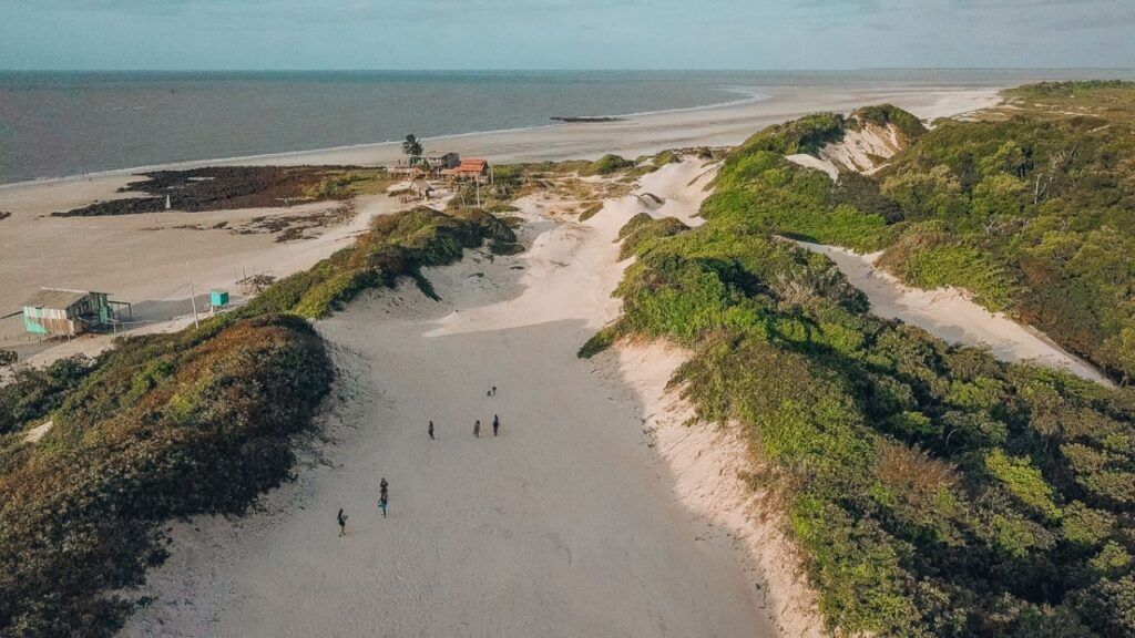 Vista aérea de una playa con algunas personas caminando, un edificio pequeño y vegetación densa.