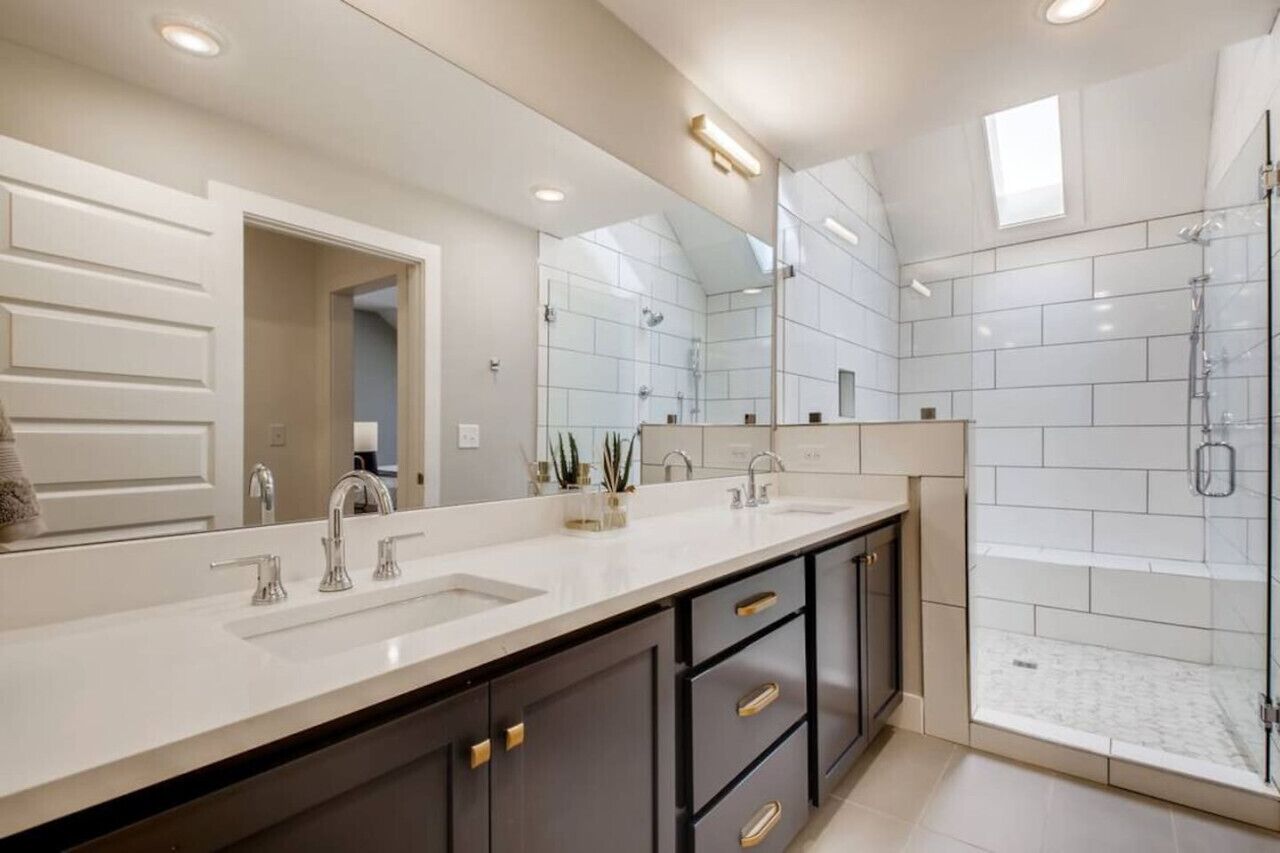 A modern bathroom with a large mirror, two sinks, dark cabinets, and a glass-enclosed shower area.