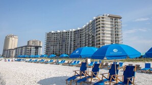 Ein Strandhotel mit mehreren Etagen, blauen Sonnenschirmen und Liegestühlen.