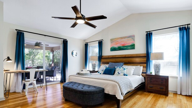 Une chambre à coucher comprenant un lit, un bureau, une chaise, une fenêtre avec des rideaux, un ventilateur de plafond et un tableau accroché au mur.