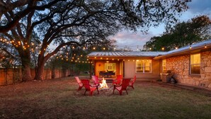 Une maison en pierre avec un foyer et des chaises rouges dans la cour, entourée d’arbres et illuminée de guirlandes lumineuses.