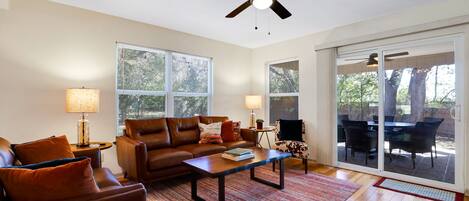 Un salon avec un canapé en cuir marron, une table basse en bois et un ventilateur de plafond. Il y a deux fenêtres et une porte-fenêtre coulissante qui donne sur un patio extérieur aménagé avec une table et des chaises.