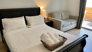 A neatly made bed with white linens, a bedside table with a lamp, a wicker basket with towels, and a grey sofa in a hotel room.