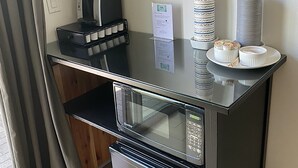 A kitchenette with a coffee maker, stacked paper cups, a microwave, and a small plate with a dessert.