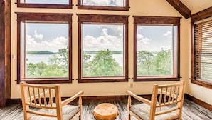 A room with large windows offering views of a lake and trees. There are two wooden chairs and a small round table.